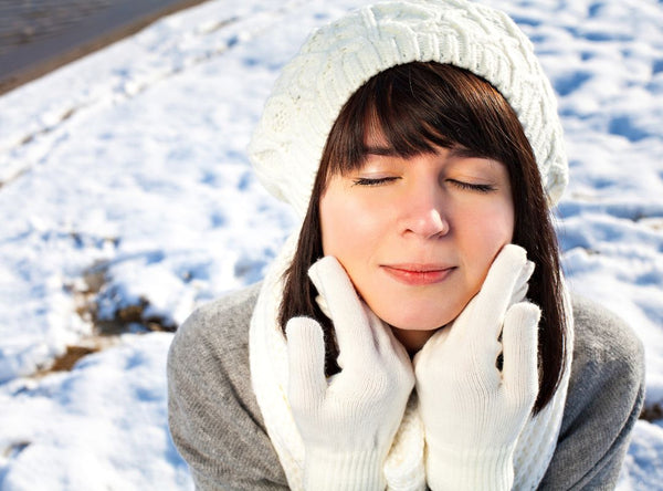 There is a woman with bangs wearing a white beanie, gloves, and scarf. She is outside in the snow and holding the back of her hands to her face.